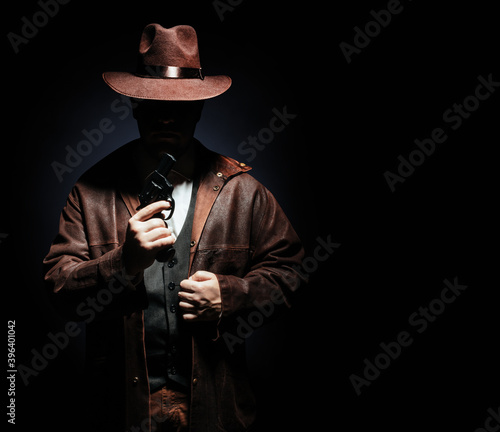 Photo of a shaded detective in jacket and hat holding a revolver gun on black background.