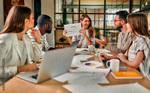 Young beautiful business woman in glasses shows a graph on paper to a group of colleagues sitting at a table with laptops in a modern office.
