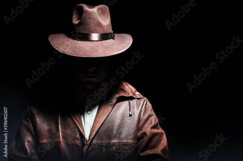 Photo of a shaded detective in jacket and hat on black background.