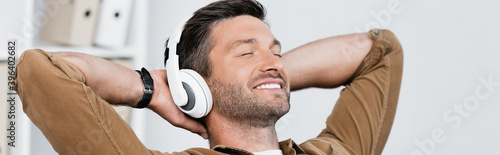 Smiling businessman in headphones, with hands behind head and listening to music on blurred background, banner photo