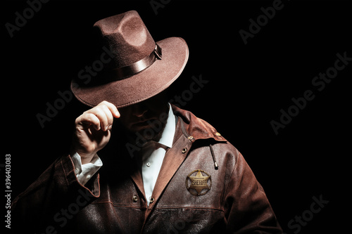 Billede på lærred Photo of a shaded sheriff officer with badge in jacket putting on cowboy hat on black background