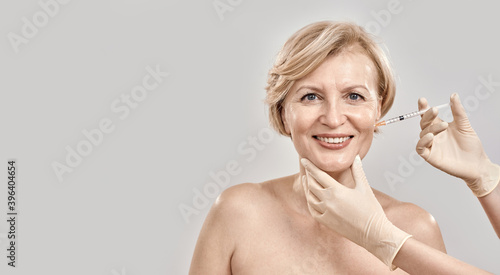 Portrait of beautiful naked middle aged woman smiling at camera while she gets injection in her face, posing isolated against grey background