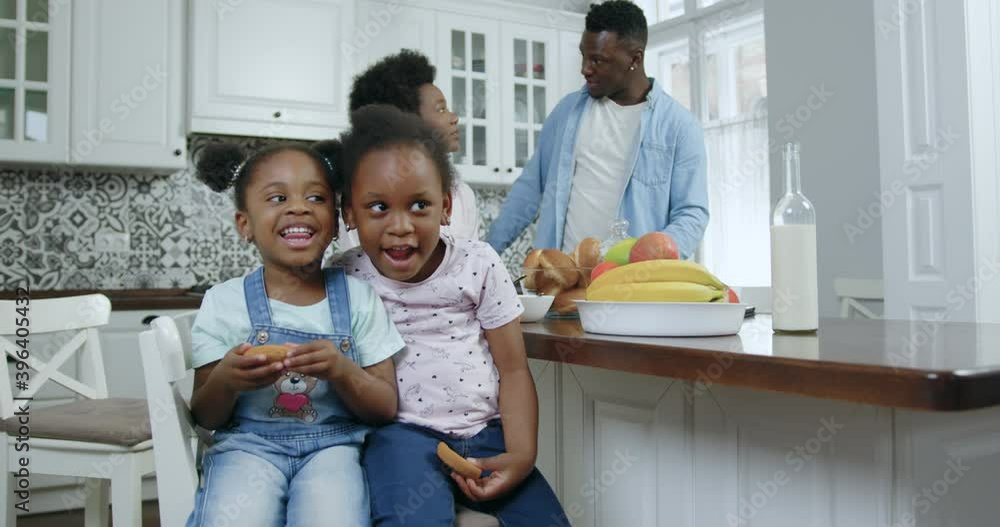 Attractive positive little two black-skinned girls making funny grimaces on camera while sitting near the kitchen table near their parents,close up,4k Attractive positive little two black-skinned girls making funny grimaces on camera while sitting near the kitchen table near their parents,close up,4k