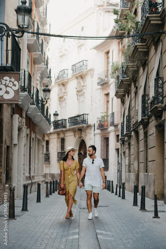 A girl in a hat and a yellow dress with a plunging neckline and her boyfriend with a beard are walking and staring at each other in old Spain town. A couple of tourists on a date in Valencia.