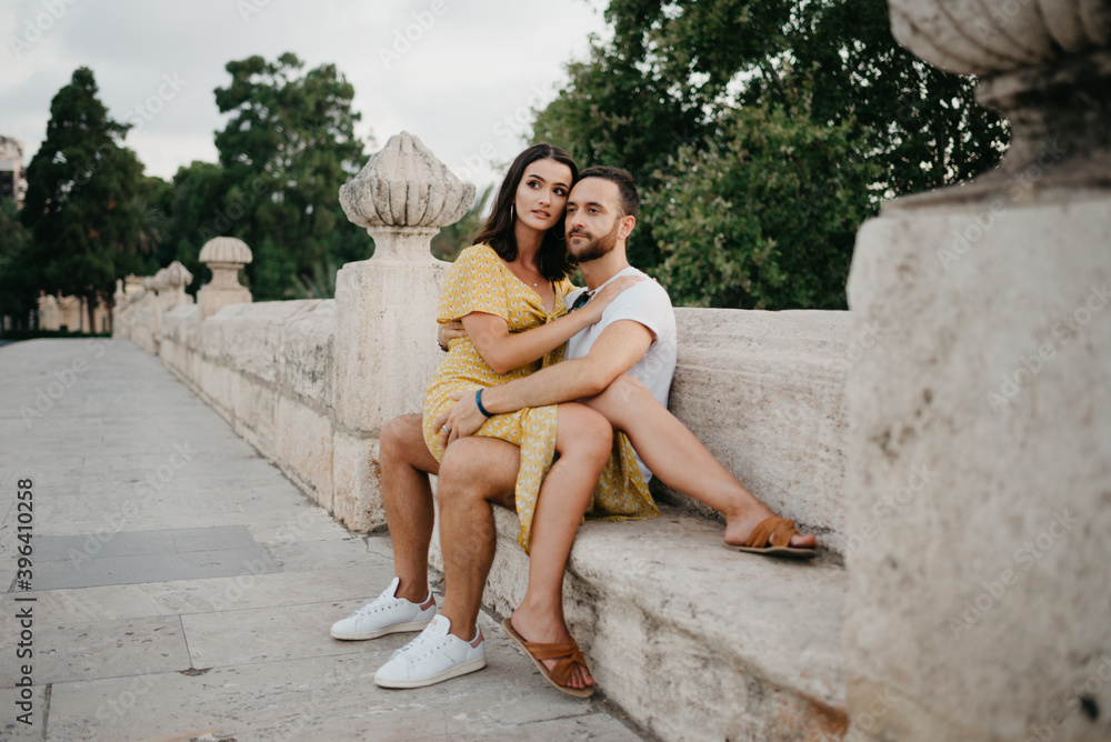 A brunette girl in a yellow dress who is sitting on the legs of her boyfriend and hugging him on the ancient bridge in old Spain town. A couple of tourists on a date in Valencia.