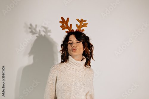 Pretty happy young woman with curly hair wearing white sweater and christmas headpear sending a kiss over isolated background