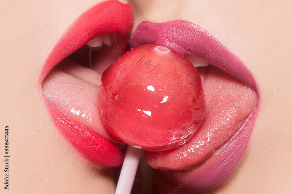 Sensual female mouth lick. Lesbian oral pleasure. Closeup view of two sexy  sensual bright female lips with pink and purple lipgloss holding in mouth  and licking delicious round red lollipop candy. Stock