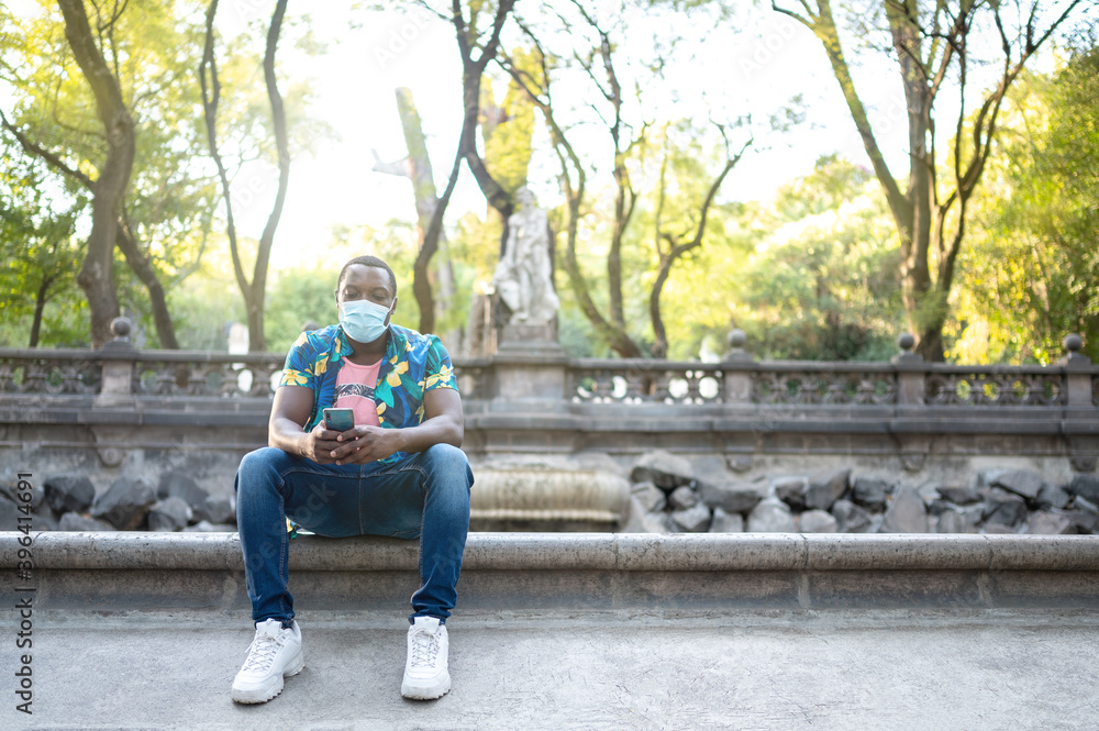 Obraz premium A young black man with a mask in the covid-19 pandemic season for protection. African American man with cell phone. Portrait of a young african man with a face mask in park.