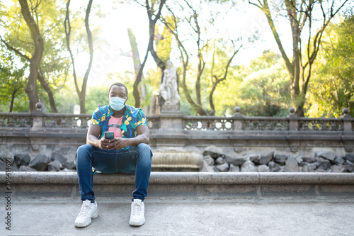 A young black man with a mask in the covid-19 pandemic season for protection. African American man with cell phone. Portrait of a young african man with a face mask in park. © Rodrigo Lucentini
