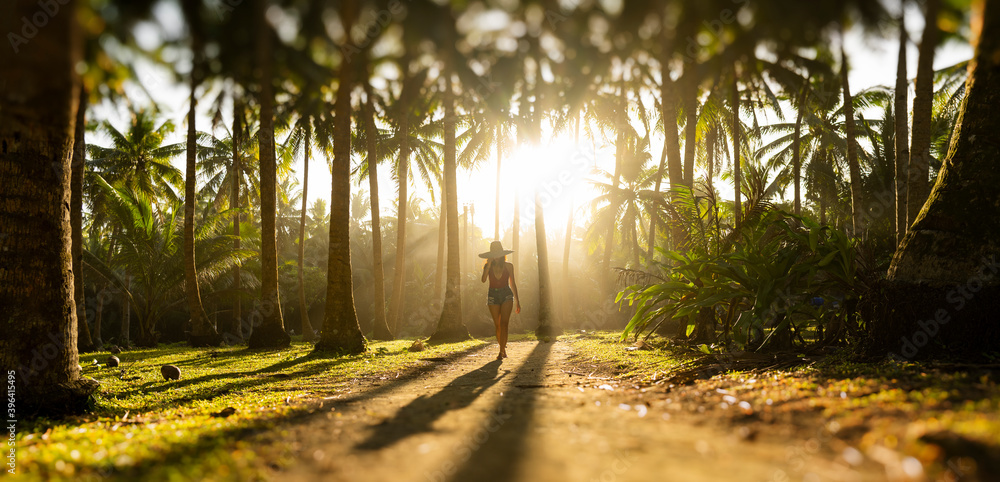 Silhouette of a beautiful girl wearing a large hat and a red bikini walking on a pathway surrounded by coconut palm trees during a stunning sunset. Siargao Island, Philippines.