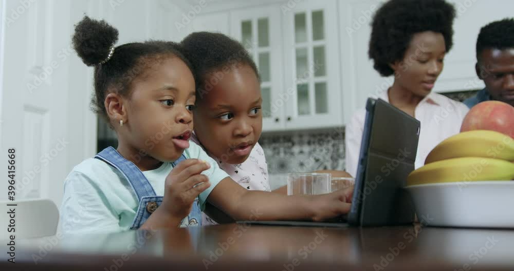 Cute interested small african american two sisters sitting at dinner table in contemporary kitchen and watching cartoons on tablet pc,4k,close up Cute interested small african american two sisters sitting at dinner table in contemporary kitchen and watching cartoons on tablet pc,4k,close up