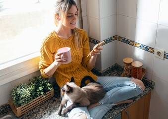 Pretty young woman with her cute cat using mobile phone while drinking coffee sitting on kitchen table at home.
