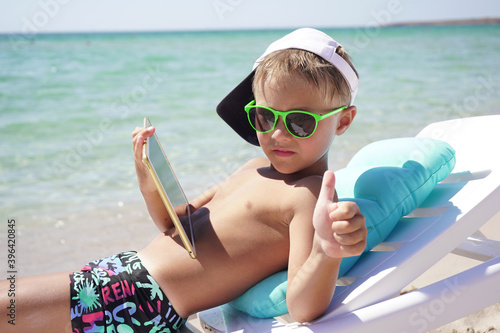 funny boy uses tablet on the beach. A child is sitting in a sun lounger on the beach against the background of the azure sea with a tablet in his hands.