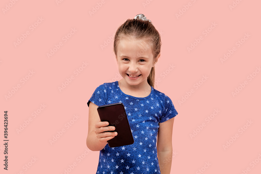 The girl looks at the smartphone screen with a smile on her face. Human emotion. In a blue dress with a small star print. Pink isolated background. The girl looks at the smartphone screen with a smile on her face. Human emotion. In a blue dress with a small star print. Pink isolated background.