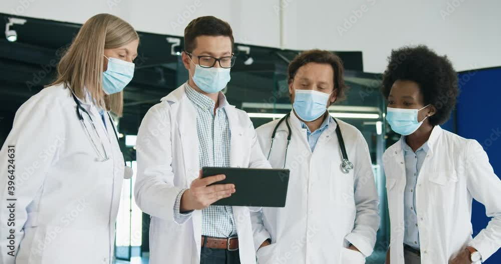 Male and female mixed-races doctors in medical mask discussing coronavirus disease while typing on tablet standing in hospital. Portrait of healthcare professionals at work. Covid 19 pandemic Male and female mixed-races doctors in medical mask discussing coronavirus disease while typing on tablet standing in hospital. Portrait of healthcare professionals at work. Covid 19 pandemic