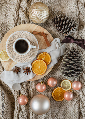 flat lay of a cup of coffee on wooden round tray on knitted beige blanket background with Christmas decorations around it: Christmas ornaments, pine cones, ribbons, dried oranges, cinnamon and anise