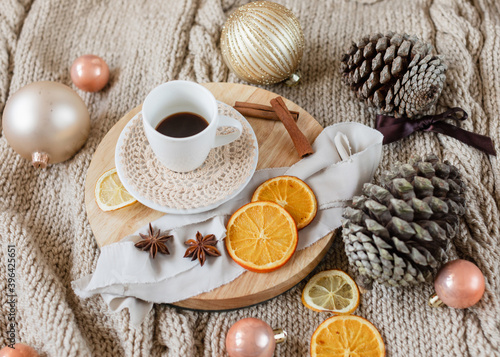 cup of coffee on wooden round tray on knitted beige blanket background with Christmas decorations around it: Christmas ornaments, pine cones, ribbons, dried oranges, cinnamon and anise