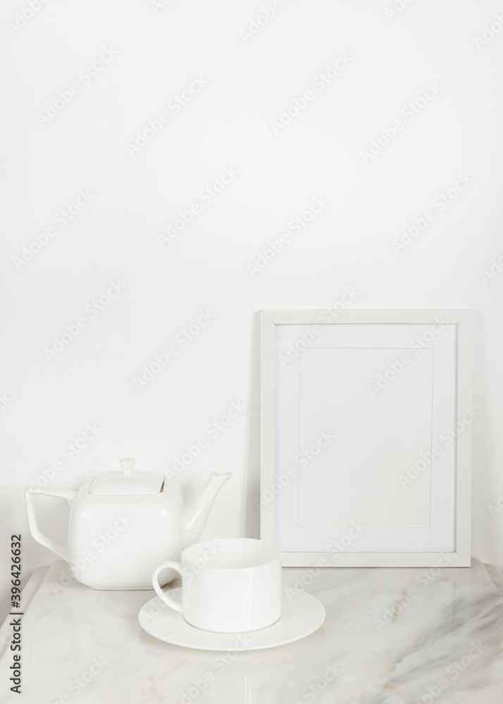 Blank white photo frame on marble surface against white wall, near a white porcelain teapot and a. white tea cup 