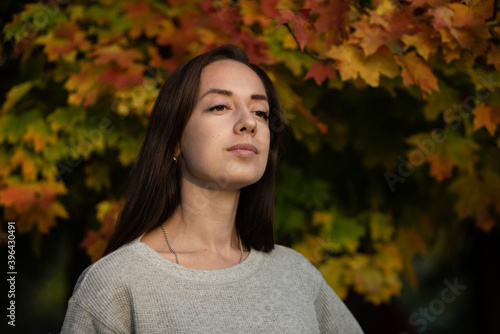 A young girl stands against the background of an autumn maple tree. He smiles slightly and looks into the distance.