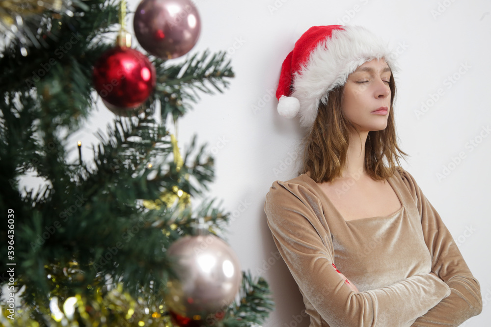Portrait of young stressed woman wearing Santa hat next to the Christmas tree at home. Winter holidays stress concept.