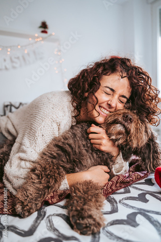 A beautiful brunette woman lying down on the bed with her Spanish water dog while hugging it. She has a very expressive face and she has a big smile. Family dog Christmas concept