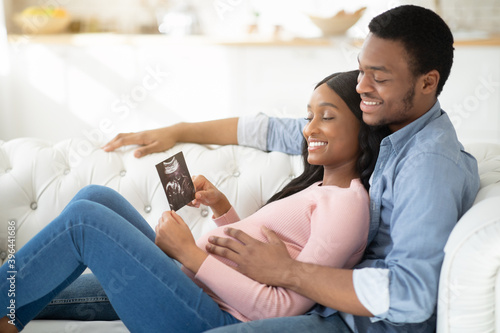 Portrait of positive black couple with ultrasound photo of their future baby cuddling on sofa at home