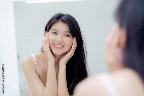 Beautiful young asian woman smiling look at mirror of checking face with skin care and cosmetic for rejuvenation and hygiene, beauty girl happy clean facial with cream or lotion and for health.