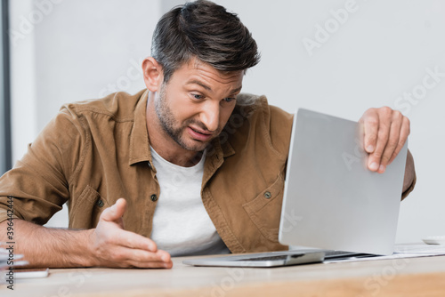 Dissatisfied businessman gesturing while looking at broken laptop at workplace on blurred foreground © LIGHTFIELD STUDIOS