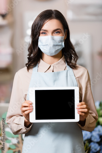 florist in medical mask holding tablet with blank screen while looking at camera