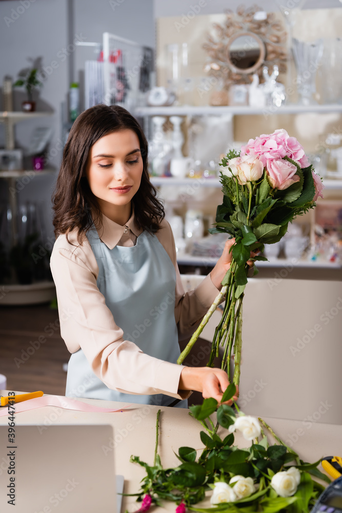 Positive female florist taking rose from desk, while arranging bouquet with blurred racks of vases on background Positive female florist taking rose from desk, while arranging bouquet with blurred racks of vases on background