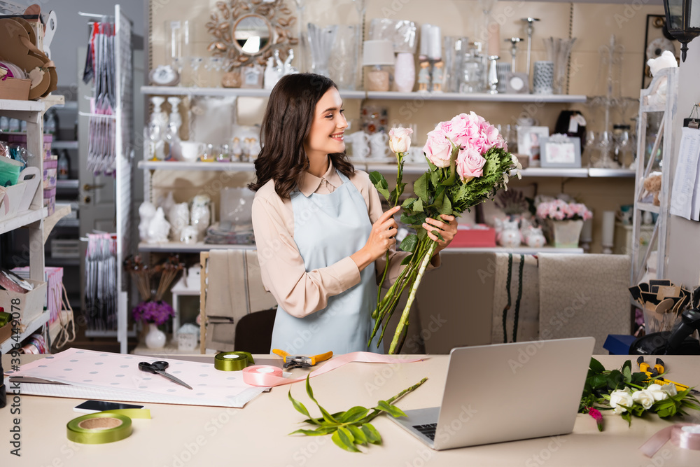 Happy florist composing bouquet with flowers, while standing near workplace with tools and laptop in flower shop