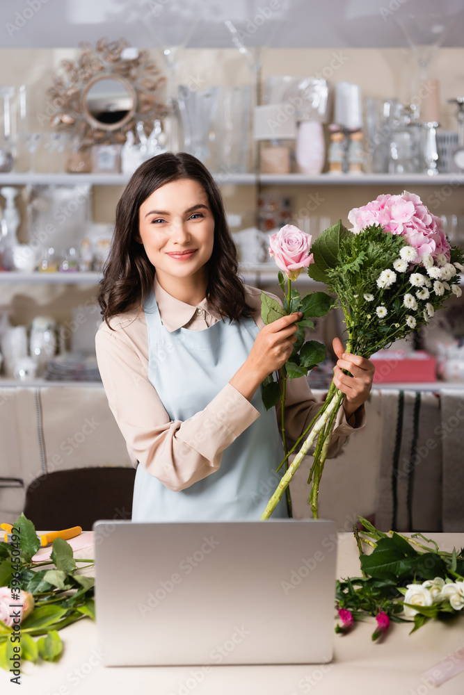 Smiling female florist looking at camera while composing bouquet near laptop on desk with blurred racks on background