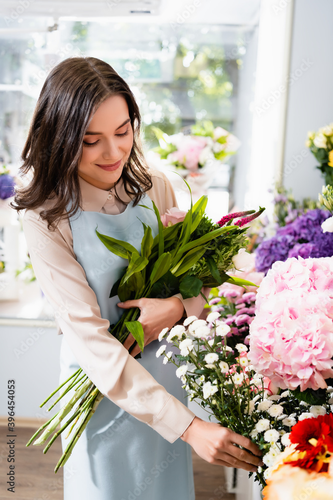 Smiling florist taking branch of chrysanthemum, while gathering bouquet near range of flowers with on blurred background