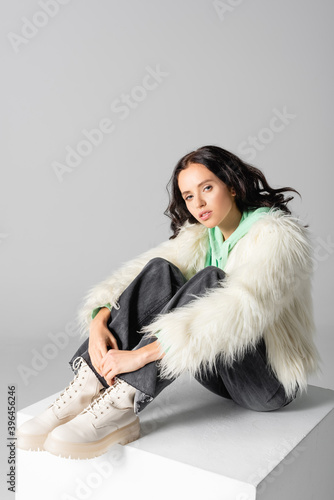  young woman in faux fur jacket posing on cube on white background