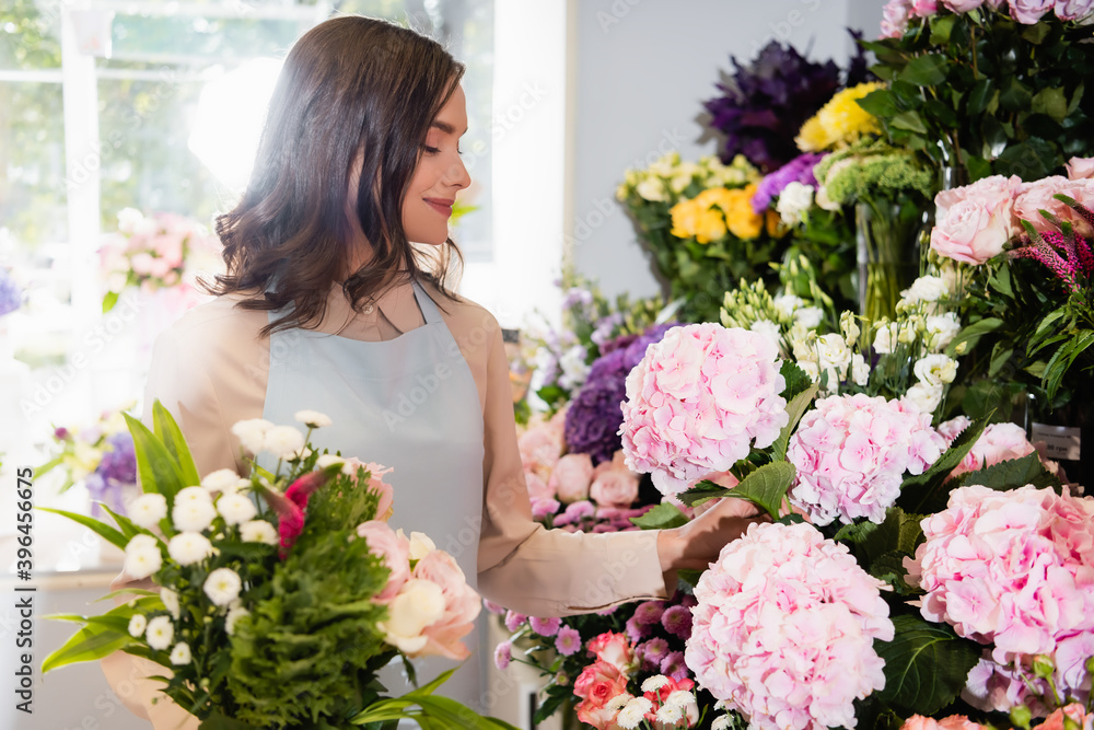 Smiling florist with bouquet taking hydrangea in flower shop with blurred window on background Smiling florist with bouquet taking hydrangea in flower shop with blurred window on background