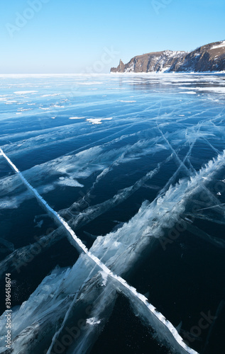 Winter landscape of frozen lake Baikal on February sunny day. Beautiful blue transparent ice with lines of cracks near the coast of Olkhon Island. Famous Hoboy Cape on the distance. Natural background