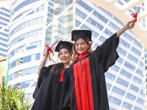 happy graduated women in graduation gowns, holding diplomas, smiling at camera. City building background. Education, successful and friendship concept.