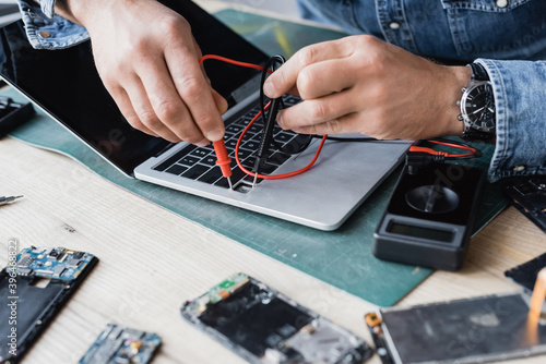 Cropped view of repairman holding sensors of multimeter on broken keyboard of laptop with blurred disassembled phones on foreground