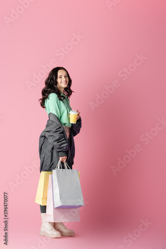  brunette young woman in casual winter outfit with shopping bags and coffee to go on pink background