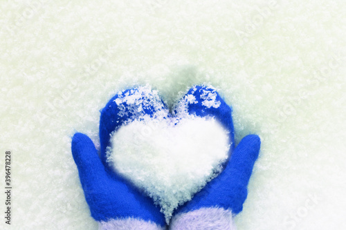 hands in knitted mittens with a heart made of snow on a winter day. Snowy heart in hands. Human hands in warm blue mittens with a snowy heart on a background of snow. Love winter christmas or romantic