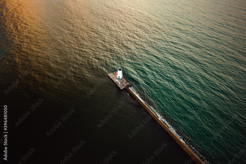 Incredible look down aerial photograph of the Ludington Breakwater Lighthouse sitting at the end of a concrete pier separating dark water from green or blue water lit up by the sun as it begins to set