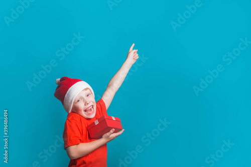 A European boy in a Santa Claus costume points to an empty space for the text. Blue background in the Studio. Copy space. The child smiles and holds a gift box in his hands. Cute boy gives a gift