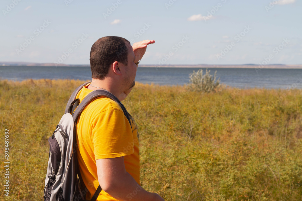 Fototapeta premium A man in an orange t-shirt with a backpack looks out to sea. The traveller shaded his eyes with his hand.