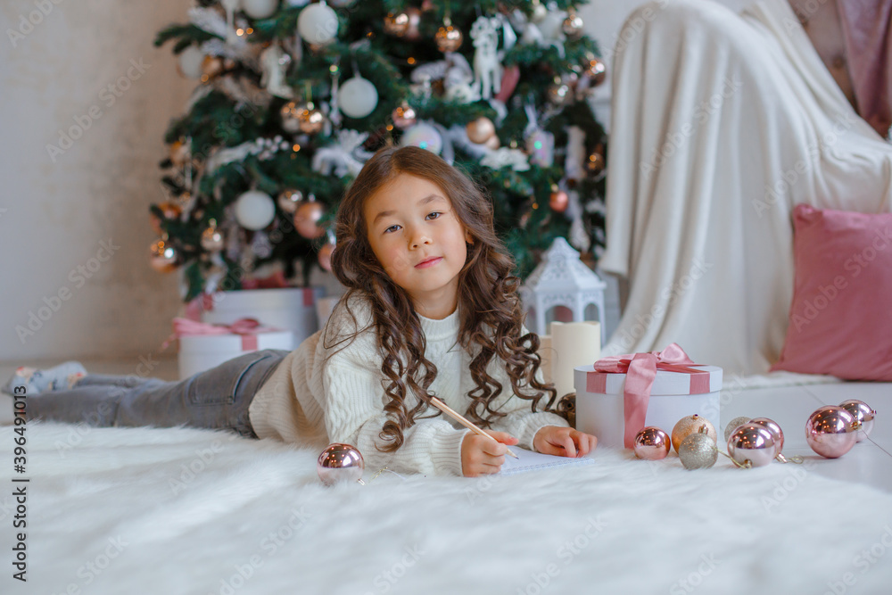 little Asian girl writes a letter to Santa Claus near the Christmas tree smiles Christmas, new year little Asian girl writes a letter to Santa Claus near the Christmas tree smiles Christmas, new year
