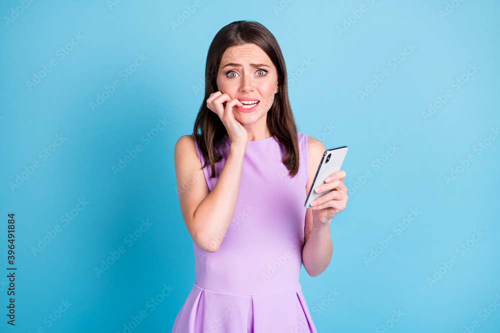 Photo of guilty scared young woman hold smartphone comment mistake bite nail isolated on blue color background Photo of guilty scared young woman hold smartphone comment mistake bite nail isolated on blue color background