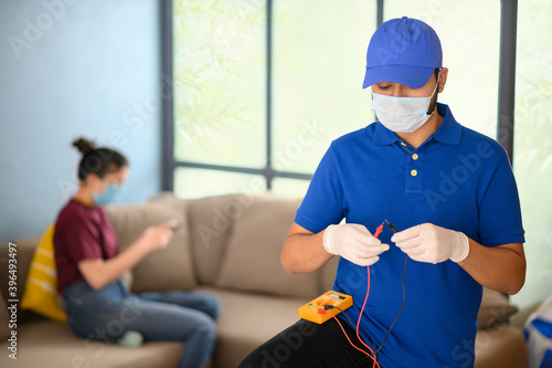 Electrician testing the electricity with electro meter with a woman sitting on the sofa in background	 photo