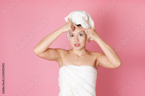 mock up a young woman in a towel squeezing out a pimple on her forehead or doing a facial massage isolated on a pink background.