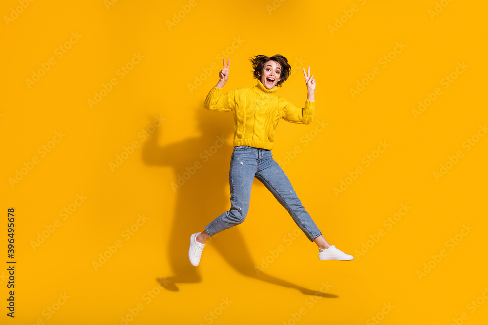 Full length body size photo of jumping high girl showing v-sign peace with both hands isolated on bright yellow color background