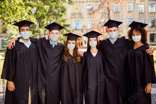 Photo of six successful young graduates wear black robes hats masks finished their education outside