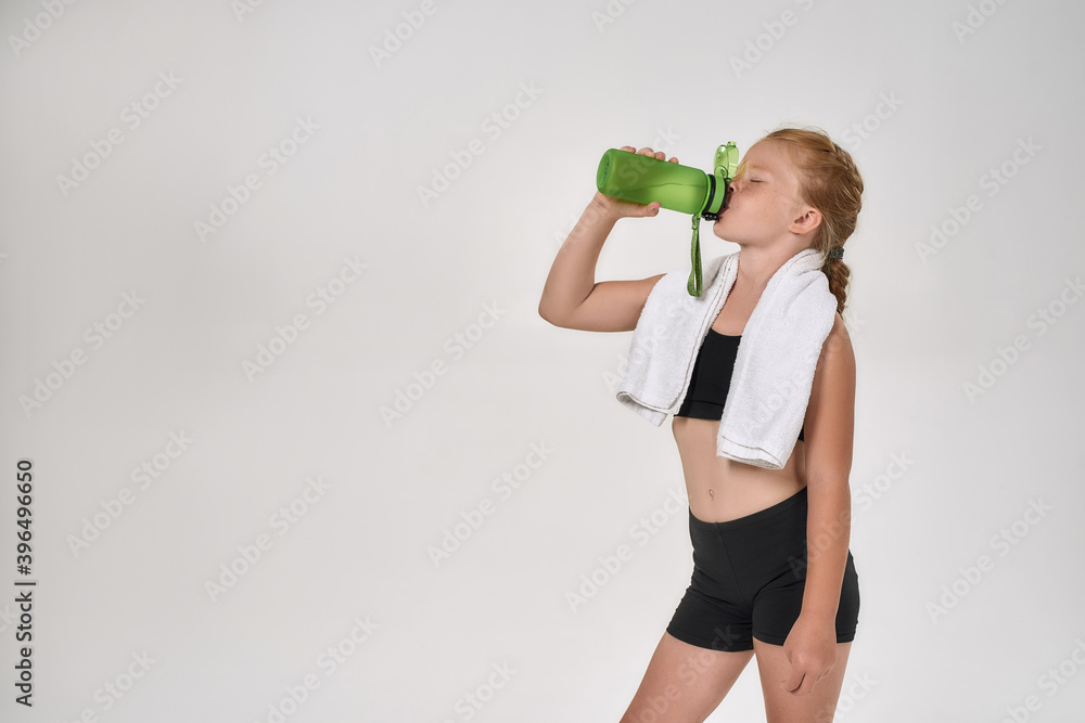 Pretty little red haired sportive girl in sportswear drinking water from the bottle, posing with white towel around her neck isolated over white background Pretty little red haired sportive girl in sportswear drinking water from the bottle, posing with white towel around her neck isolated over white background